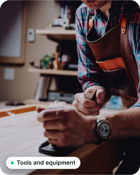 A joiner in a checked shirt and leather apron sanding a timber board at a workbench